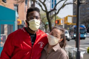 Two people wearing masks outdoors, arms around each other, looking at the camera, outdoors. 