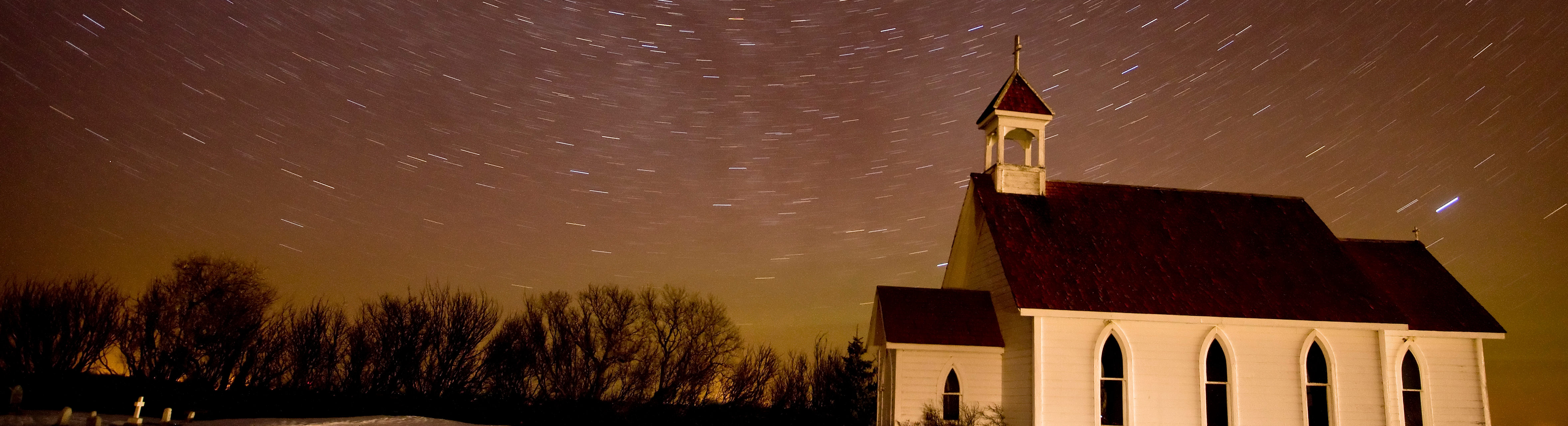 White country church with trees along the bottom and night sky with star trails above. 