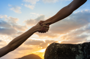 Two hands and forearms, stretching out diagonally to clasp hands, with a sunrise in the background. 