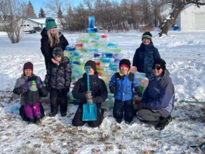 Kids, adults and a baby sit in all their winter gear in front of a lovely rainbow ice wall in a snowy field.